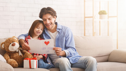 Lovely little girl and her dad happily celebrating Father Day together, sitting on sofa at home, reading greeting card, copy space