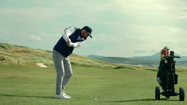 Male golfer hitting approach shot on green manicured fairway of Ireland links golf course on perfect picturesque blue sky day next to his golf bag in slow motion