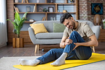 Contorted in pain, a young Indian man sits on an exercise mat at home and holds his leg with his...