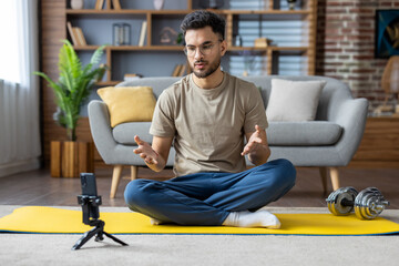 Fototapeta premium Young Indian male trainer and instructor sitting at home on mat in front of phone and giving online lecture and training