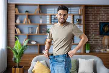 Portrait of a young smiling Indian man standing at home in front of the camera wearing jeans and showing the result of diet, exercise and weight loss