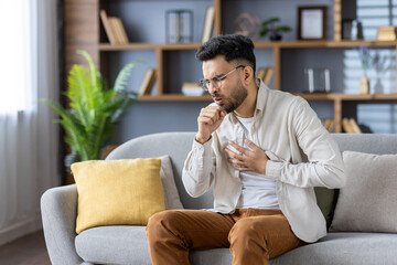 Muslim young man sitting on the sofa at home, holding his chest with his hand, feeling severe pain,...