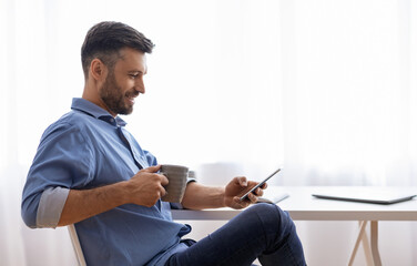 Break In Work. Relaxed Male Freelancer Using Smartphone And Drinking Coffee While Sitting At Desk...