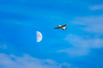 Small plane flying near a crescent moon on a blue sky with clouds.