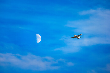 Small plane flying near a crescent moon on a blue sky with clouds.