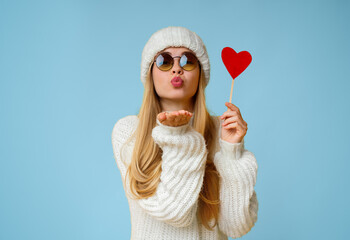 Lovely girl in winter hat and sunglasses holding red heart card and sending flying kiss, yellow...
