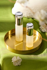 Two skincare bottles with a silver cap are placed on a golden tray surrounded by a few white flowers and a soft, green bedspread with sunlight, suitable for advertising beauty care products.