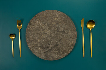Top view of brown marble plate, gold cutlery on dark green background. Food presentation, stone tray. Table setting, copy space, flat lay.