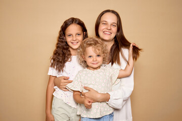 Caucasian cheerful mom older daughter and newborn boy cuddling together indoors isolated over beige background sharing happy moments and positive feelings