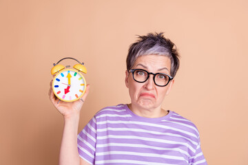 Photo of sad upset mature woman wear striped stylish clothes hold alarm clock isolated on beige color background