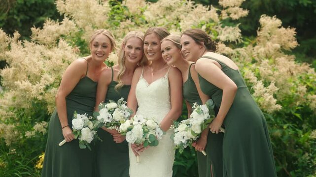 Bride and bridesmaids lean in heads together holding bouquets in garden setting