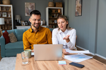 Obraz premium Working at home. Young freelance couple work from home office on laptop computer having video call with clients, showing statistics of business improvement with sales graphic printed on paper.