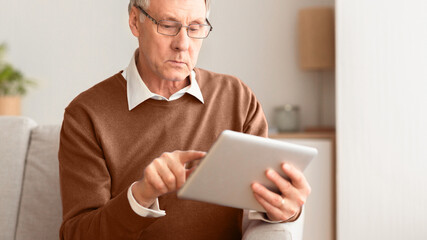 Serious Senior Gentleman Using Tablet Computer Reading News Sitting On Sofa At Home. Selective Focus, Panorama, Cropped