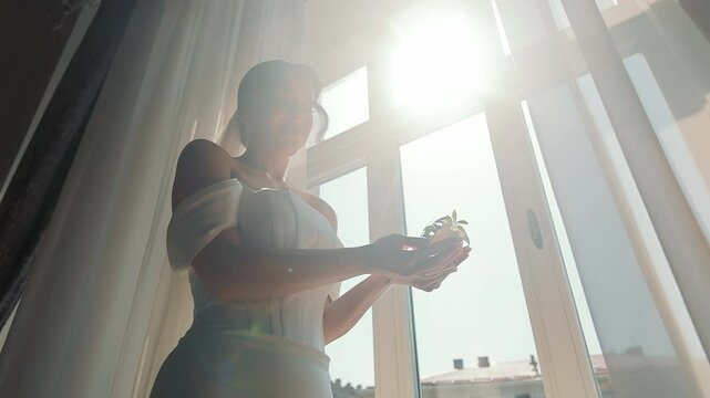 Bride Holding a Buttonhole in a Sunlit Room. A bride dressed in an elegant white gown holds a buttonhole in her hands as she stands by a large window.
