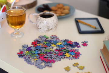 Colorful jigsaw puzzle pieces, books, tablet, glasses, cookies, berries, cup of tea or coffee, glass of juice and lit candles on the table. Hygge in pastel colors. Selective focus.