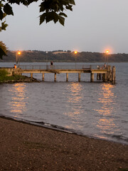 A tranquil scene of Bracciano Lake in Trevignano Romano as twilight falls. The pier lights cast warm reflections onto the lake’s surface, while a few people relax on the pier, enjoying the evening.