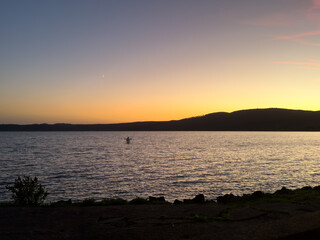 A stunning sunset over Bracciano Lake with a silhouetted figure standing on a paddleboard. The tranquil scene showcases calm waters, fading daylight, and a crescent moon rising over distant hills.