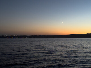 A crescent moon above Bracciano Lake in Trevignano Romano, Italy, and distant lights on the horizon under the fading dusk sky.