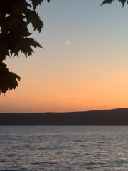 A serene moon rises over Bracciano Lake at dusk in Trevignano Romano, Italy, with calm waters framed by tree branches.