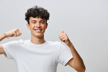 Smiling young man showing excitement with thumbs up gesture in a neutral grey background, wearing a...