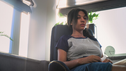 Depressed teenager sits in chair during therapy with psychotherapist in dim psychological office....