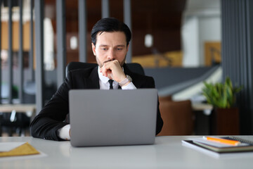 Fototapeta premium A man in a suit is sitting at a desk with a laptop open in front of him. He is deep in thought, possibly working on a project or dealing with a difficult problem. Concept of focus and concentration