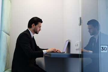 A man in a suit is typing on a laptop in a cubicle. The cubicle is numbered 30