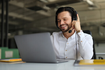 A man wearing a headset is smiling while using a laptop. He is in a professional setting, possibly working on a project or attending a meeting. Concept of productivity and focus