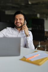 A man is talking on his cell phone while sitting at a desk. He is smiling and he is enjoying the...