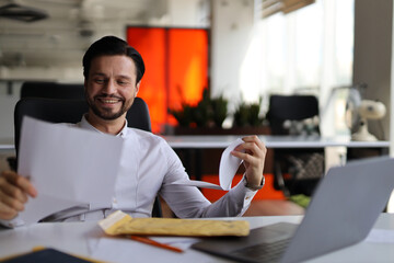 A man is sitting at a desk with a laptop and a piece of paper. He is smiling and holding the paper in his hand. The scene suggests a positive and productive atmosphere