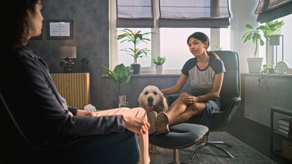 African American teen girl sits in chair, listens to psychologist, pets golden retriever dog during counseling session. Female therapist talks to young patient. Psychology, mental health. Slow Motion.