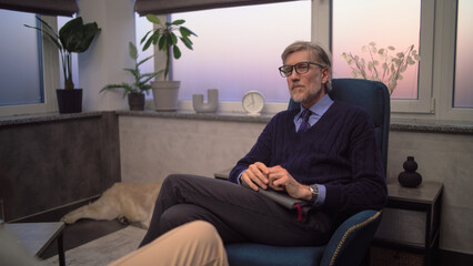 Mature psychotherapist sits in chair, listens to patient and asks questions during therapy in...