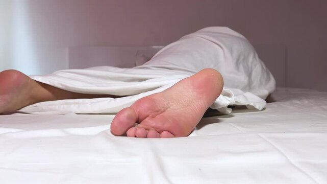 Horizontal 4K video. White adult woman sleeps in hotel room on light bed under blanket. Female leg close up. Toes. Concept of dream, sleep, relax, skin care, tourism, spa, lazy, rest. Foot are moving.