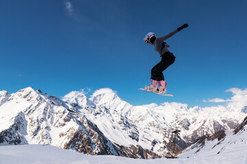 snowboarder jumps from a big air with a board grab, a nose or tail trick in rotation, against the backdrop of snow-capped mountains and the bright sun