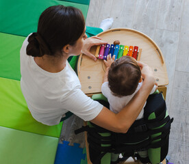 Little boy with damage to CNS participates in sensory and physical therapy standing verticalizer and using xylophone while guided by a caregiver in rehabilitation room