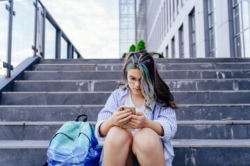 Preteen girl with blue colorful hairstyle primary school pupil sitting with backpack with emotional problems, victim of bullying in schoolyard.