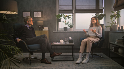 Obraz premium Woman sits in chair, talks and shares positive emotions with psychologist on counseling therapy session. Mature professional therapist listens to female patient, provides psychotherapy. Psychology.