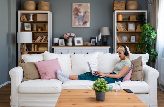 Young happy freelance worker or university student woman sitting work at home on sofa working on laptop, while listening podcast or listen the music, favorite song, enjoying freedom and independence.