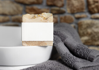 Soap bar with blank label on vessel sink near grey towels against stone wall close up, mockup