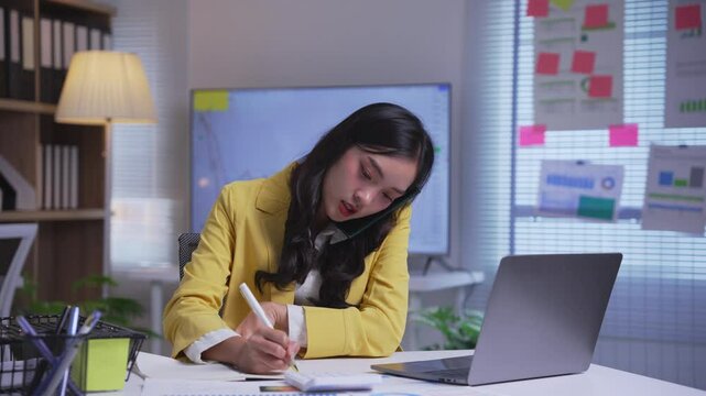 Young professional woman is multitasking in her office, balancing a phone call while diligently taking notes, showcasing the demands of a modern workplace