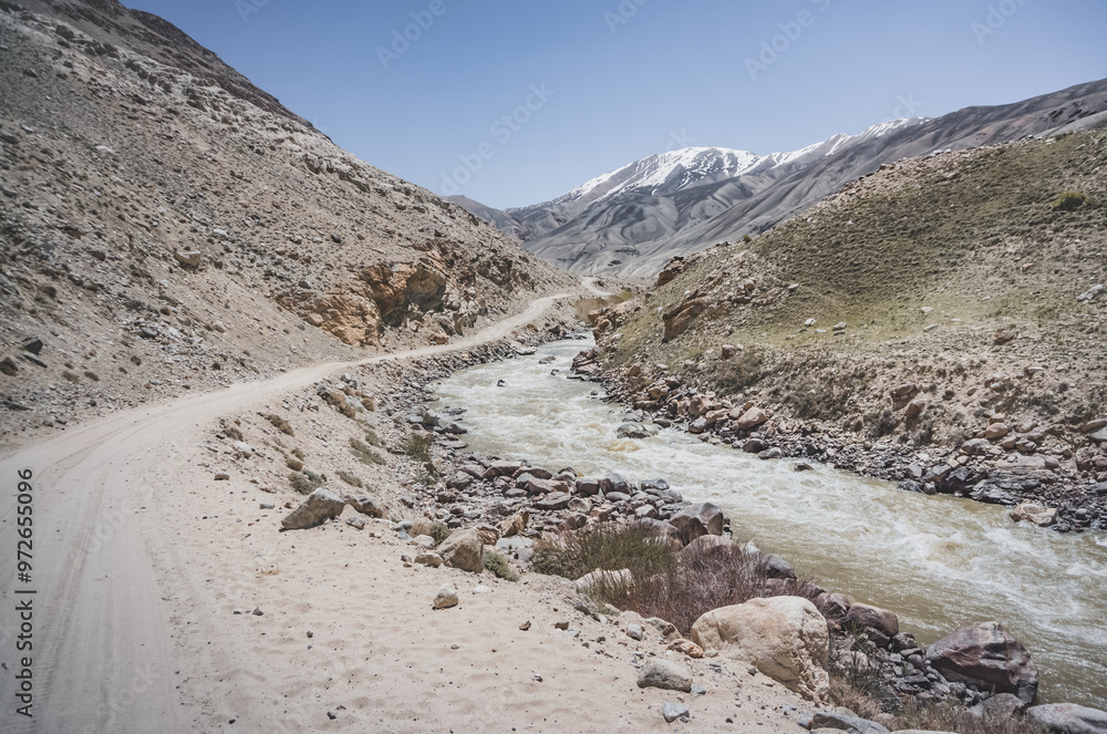 Sticker Dusty road of the Pamir Highway winds and twists in the valley of the Tien Shan Mountains in Tajikistan in the Pamirs and the Pamir River with blue clear glacial water flows in the valley