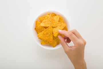 Young adult woman hand fingers taking yellow tortilla chips from white bowl on light gray table...