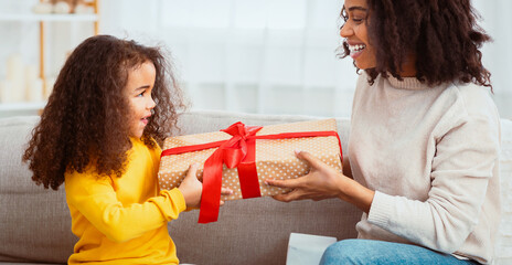 Kid's Birthday. Smiling Afro Mother Giving Daughter Wrapped Gift Congratulating On Holiday Sitting On Sofa Indoor. Panorama