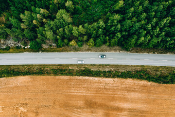 Aerial top view of asphalt road with cars between green summer woods and yellow wheat field in Finland.
