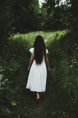 Naklejka premium A young woman in a white dress walks along a forest path. An abyss is implied, getting lost, danger lurks in the forest