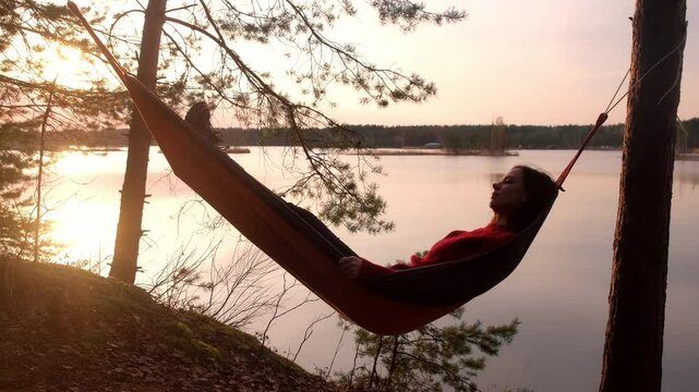 Calm relaxed woman lying in hammock in forest admiring lake nature at sunset. Resting relaxed young female in red sweater enjoying time in camping in dark woodland. Tourist, traveler, camper outdoors.