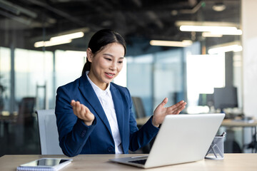 Confident Asian business woman engaging in video call in office. Smiling professional uses laptop to communicate virtual meeting. Scene conveys ideas of remote work, communication, and connection.