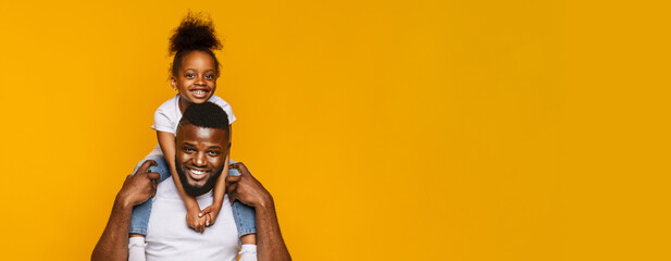 A cheerful black father carries his smiling daughter on his shoulders, both filled with laughter. They are enjoying a moment of playful bonding, highlighted by the vibrant yellow backdrop, copy space