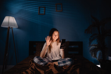 Naklejka premium young girl sitting in bed at night, having video call using smart phone