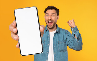 A cheerful man with a denim jacket joyfully raises a smartphone with a blank screen, showcasing an...